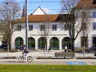 Das Dokumentationszentrum Nationalsozialismus von außen. Vor dem Gebäude stehen mehrere Bäume. Davor ist eine Straße mit Passanten u.