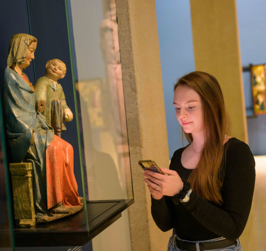 Eine Person steht vor einer Skulptur der Mutter-Kind-Gruppe und benutzt die Augustinermuseum-App auf dem Smartphone.