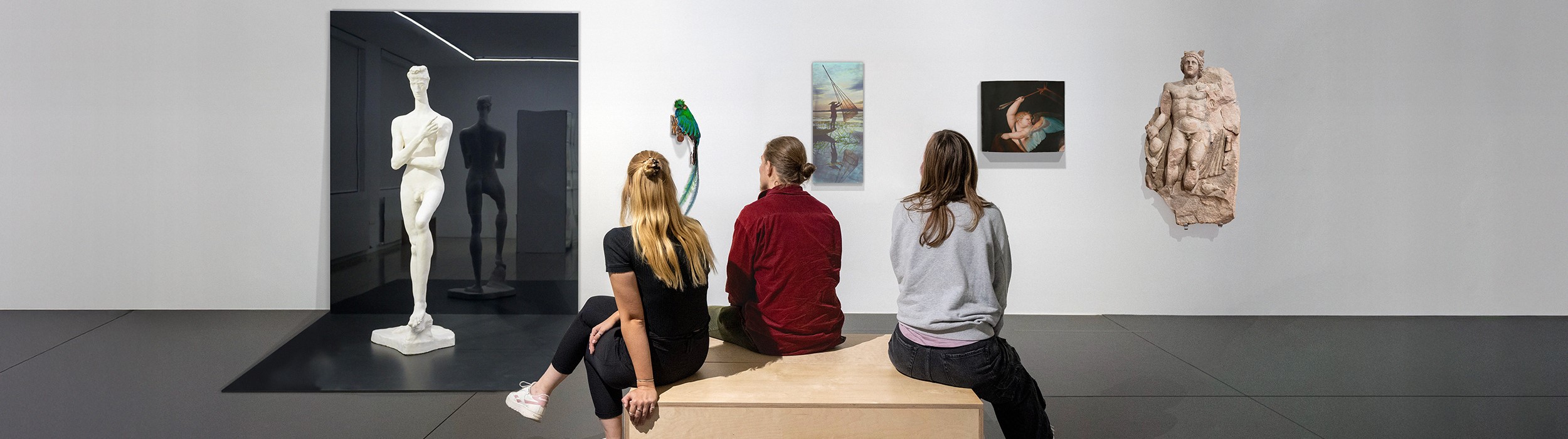 Drei Menschen sitzen auf einer Bank und betrachten eine Ausstellung, die Werke der Museen Freiburg zeigt.