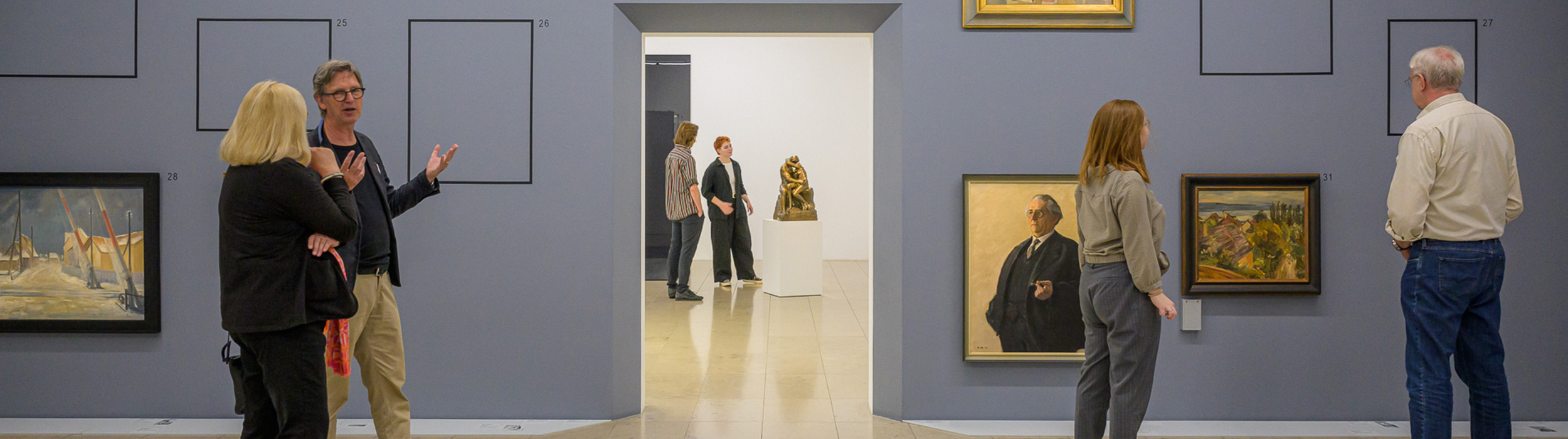 Im Vordergrund betrachten vier Personen Gemälde in einem grauen Raum und unterhalten sich. Einige Bildstellen sind frei. Durch einen Durchgang sieht man zwei Personen, die die Skulptur „Der Kuss“ von Auguste Rodin ansehen.